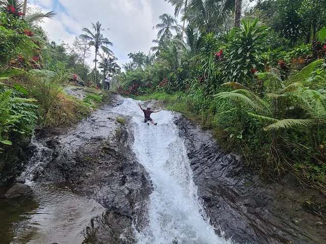 Eine Person rutscht einen kleinen natürlichen Wasserfallstrom hinunter, umgeben von tropischer Vegetation.