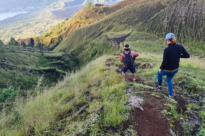 Dos personas bajan caminando por una ladera de montaña cubierta de hierba; una toca una pequeña guitarra.