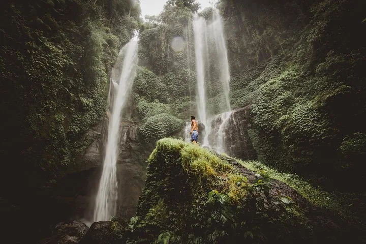 Un hombre de pie sobre una roca cubierta de musgo admira los altos chorros de la Cascada Sekumpul.